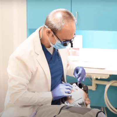 Periodontist in a white coat and blue gloves working on a patient's teeth, representing periodontal maintenance