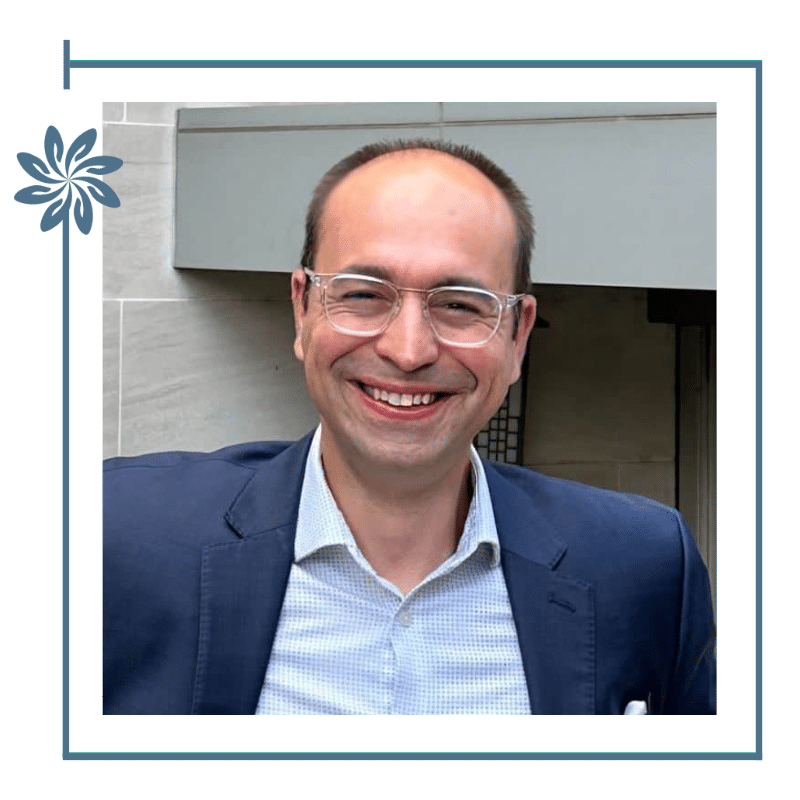 Dr. Thanos dounis smiling adult wearing glasses and a suit jacket, standing outdoors in front of a modern building facade.