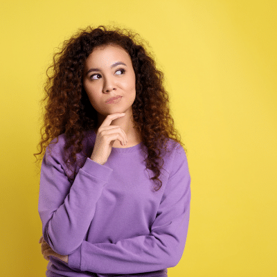 Woman with her finger on her chin looking up in thought wearing a purple sweater on a dark yellow background, representing getting a second opinion for periodontal care